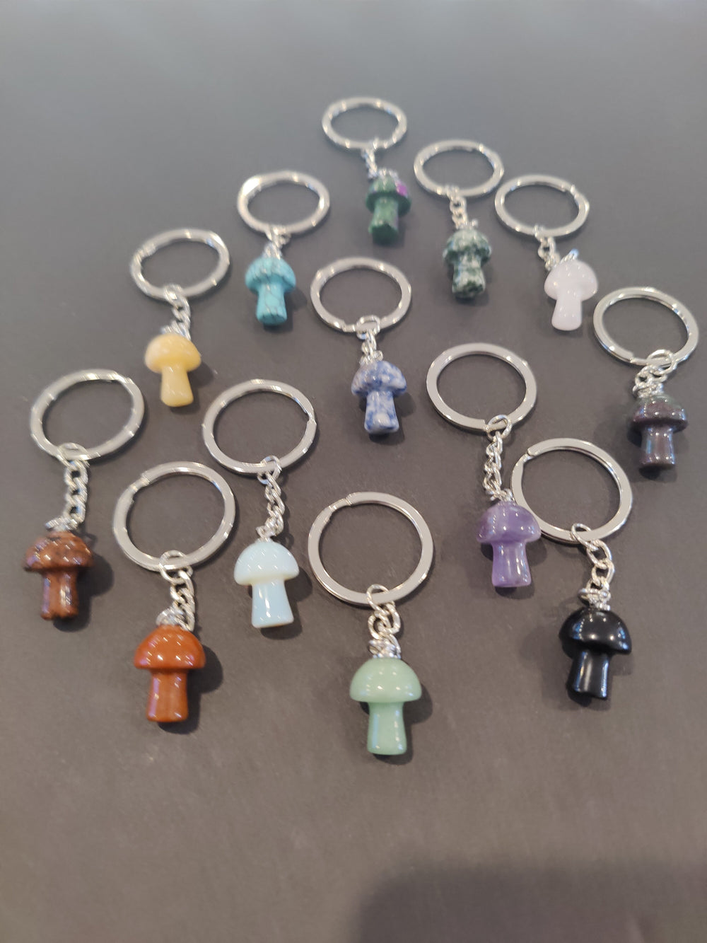 Twelve small mushroom-shaped stone keychains in various colors including orange, light blue, purple, black, and green, arranged on a gray surface.
