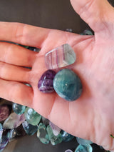 Hand holding three polished rainbow fluorite stones in purple, teal, and translucent striped colors over a surface scattered with similar stones.