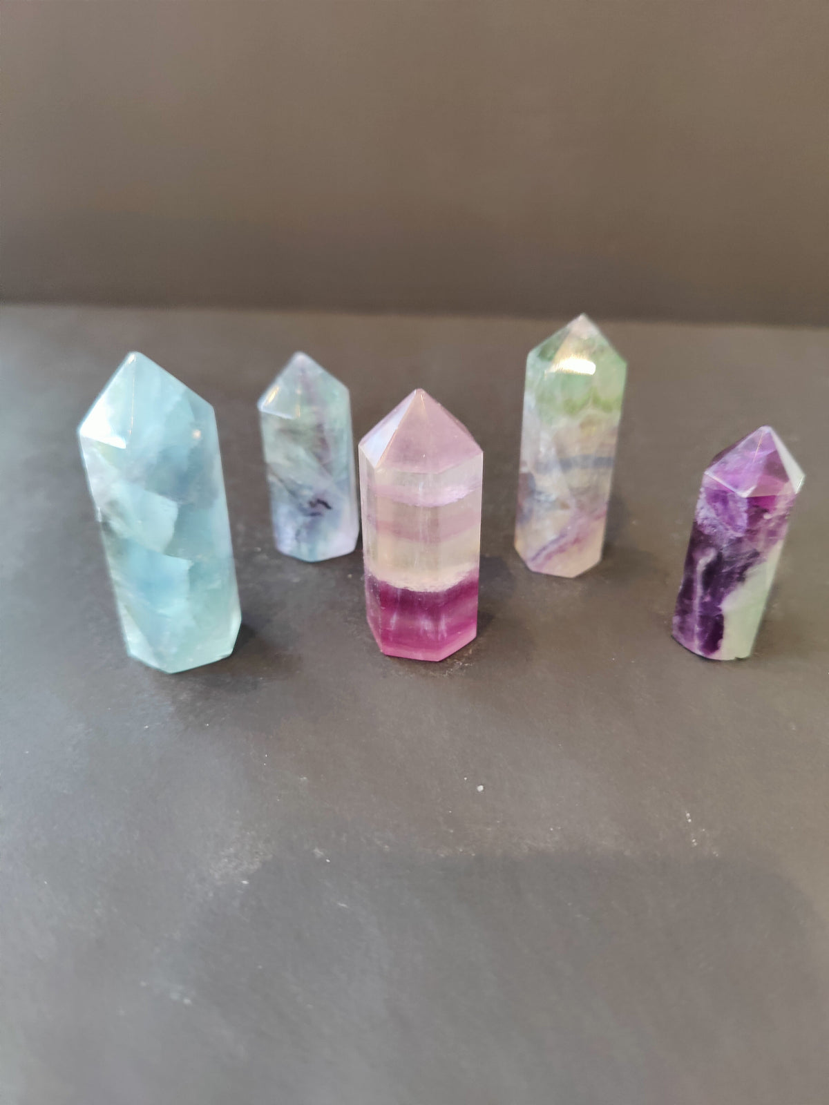 Five multi-colored fluorite crystal points in various pastel shades of blue, purple, green, and pink on a gray surface against a plain background.