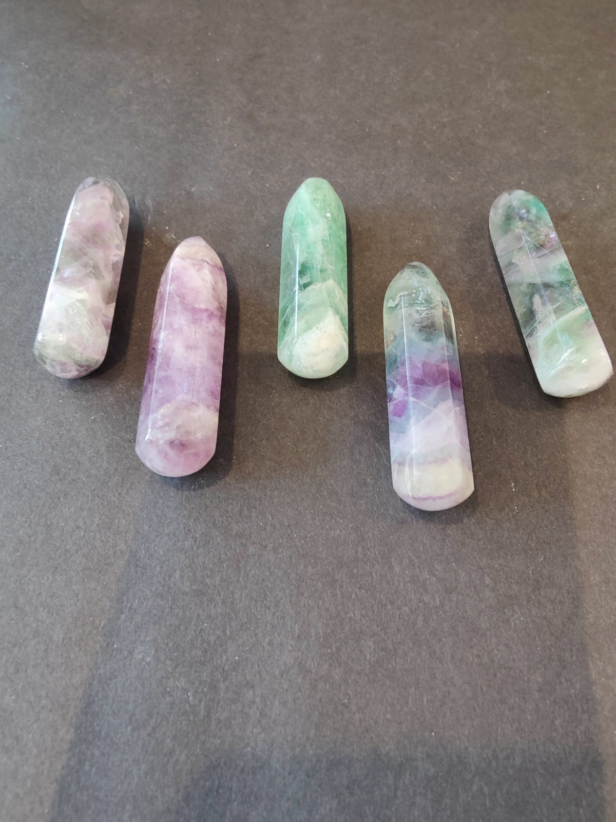 Five polished fluorite points in shades of green, purple, and white arranged on a dark textured surface with rounded and tapered shapes.