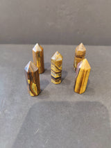 Five small polished tiger's eye crystal towers with pointed tops arranged on a gray surface, featuring rich golden-brown and dark striped patterns.