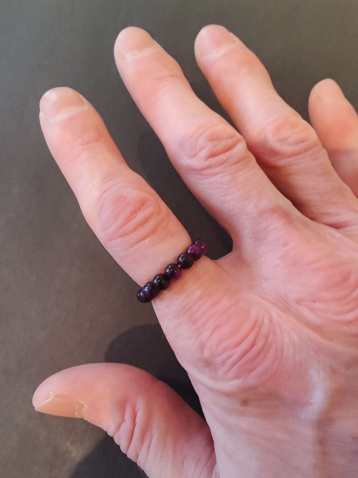 Close-up of a hand wearing a dark red beaded 4mm stone ring on the middle finger against a dark background.