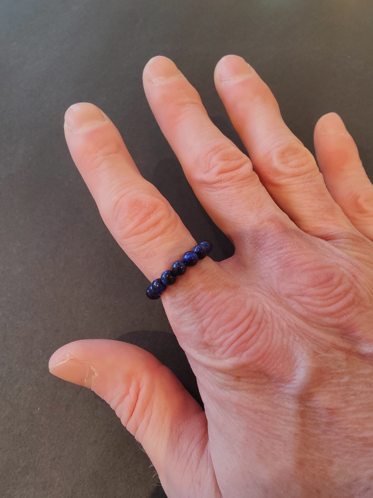 Close-up of a hand with a beaded ring of dark blue, 4mm stone beads worn on the middle finger against a dark background.
