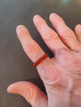 Hand wearing a delicate red beaded ring composed of small round 4mm stones on the middle finger against a dark textured background.