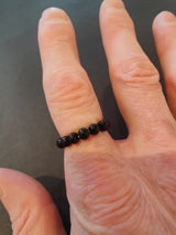 A hand wearing a beaded ring made of smooth, dark round stones on the ring finger against a plain dark background.