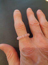 Hand wearing a delicate ring made of seven translucent light pink 4mm round beads on the middle finger, against a dark gray background.
