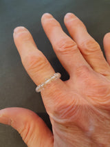 Hand with visible wrinkles wearing a translucent beaded ring on the middle finger against a dark background.