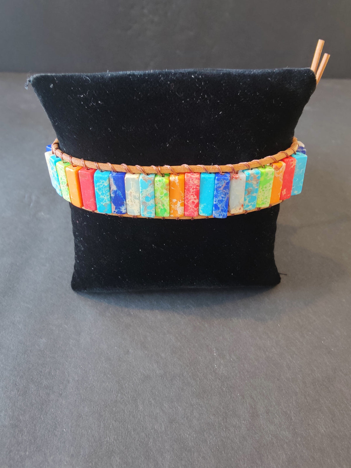 Bracelet featuring rectangular stone beads in vibrant chakra colors, woven with tan cord and displayed on a black velvet cushion.