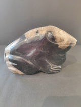 Sculpted frog figure in petrified wood, featuring smooth dark purple and natural beige tones with visible wood grain patterns.