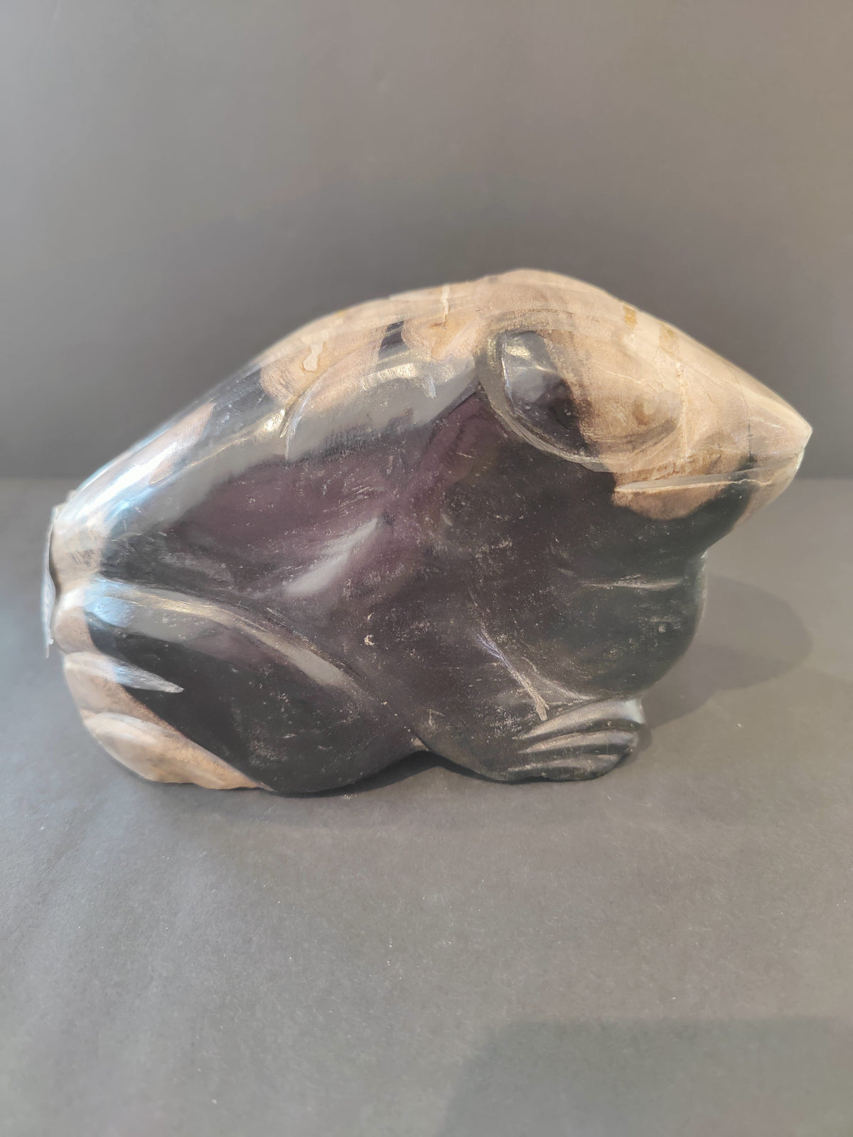 Sculpted frog figure in petrified wood, featuring smooth dark purple and natural beige tones with visible wood grain patterns.