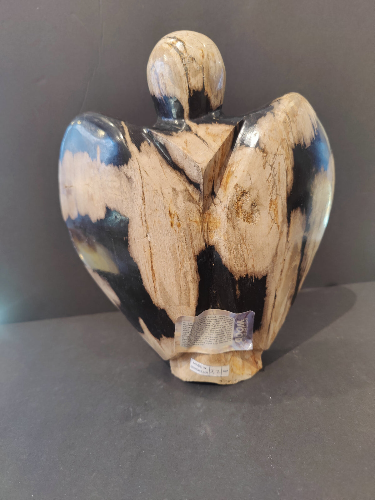 Abstract angel sculpture carved from petrified wood with light beige and dark brown marbled patterns, viewed from the back against a dark background.