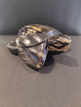Carved petrified wood turtle sculpture with dark brown and beige natural striations and smooth polished finish, shown resting on a flat surface against a gray background.