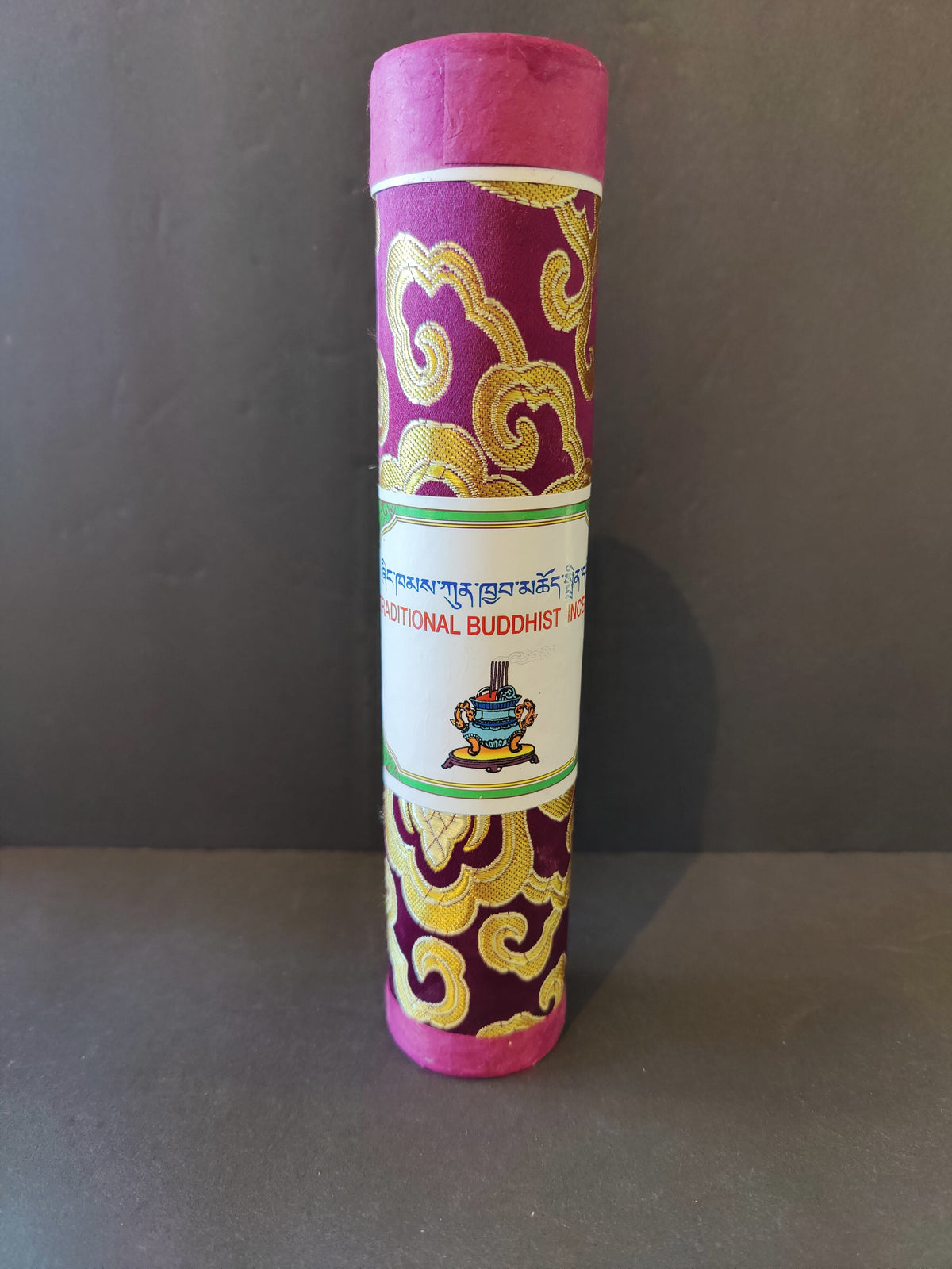 Cylindrical package wrapped in purple fabric with gold swirling patterns, featuring a white label with Tibetan script and a colorful Buddhist incense pot illustration.