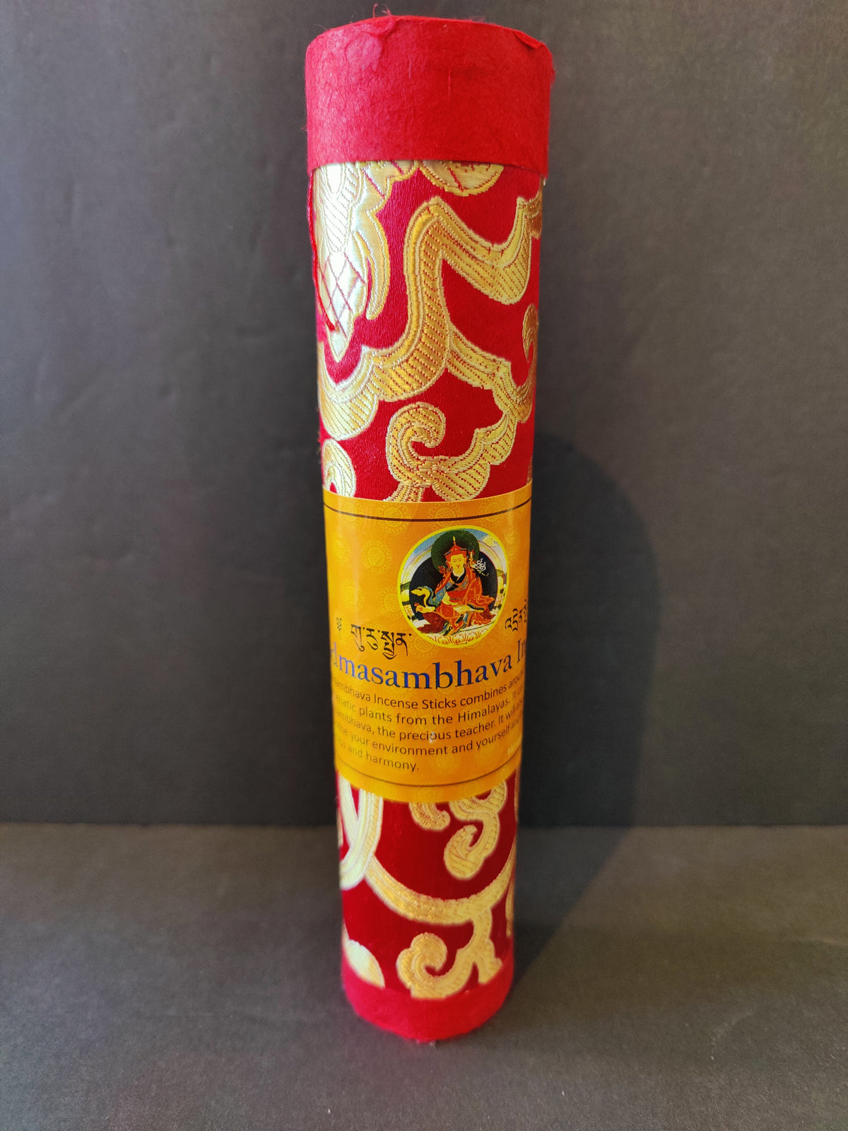 Cylindrical red and gold patterned container with a yellow label featuring a colorful Tibetan artwork, standing upright against a dark background.