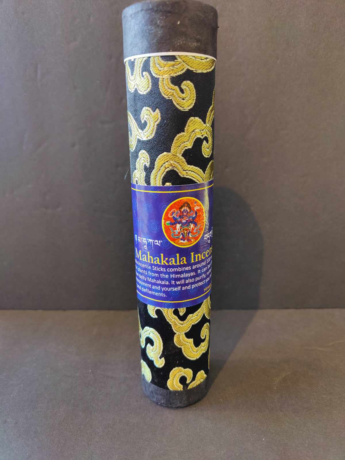 Cylindrical Tibetan incense stick bundle wrapped in black fabric with gold cloud-like patterns and a blue label featuring a red Mahakala symbol.