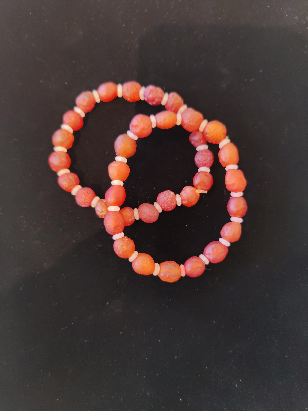 Two elastic bracelets made of round, faceted orange-red beads with small white spacer beads, arranged overlapping on a black background.
