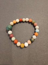 Multi-colored jasper bead bracelet featuring various shades including green, red, orange, beige, and cream, arranged in a circular pattern on a gray surface.
