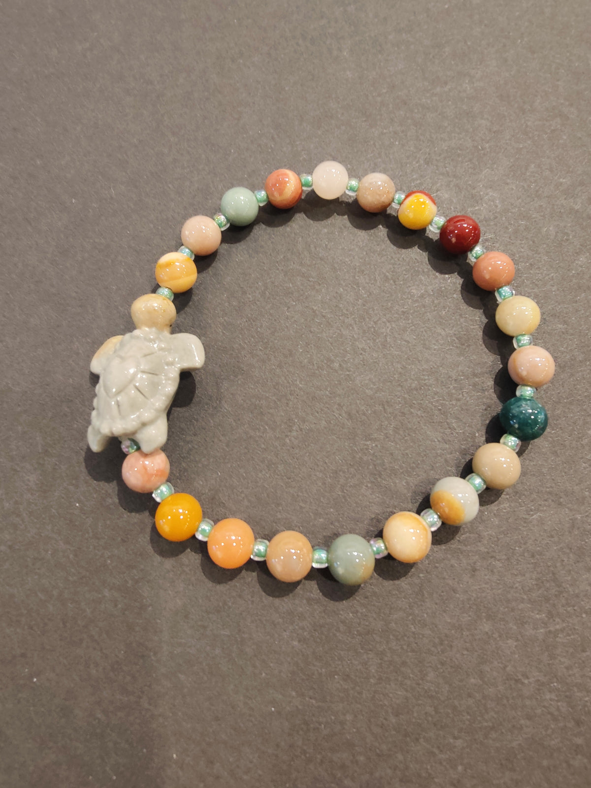 Multicolored jasper bead bracelet with a light gray turtle-shaped charm and small turquoise spacer beads on a dark gray background.
