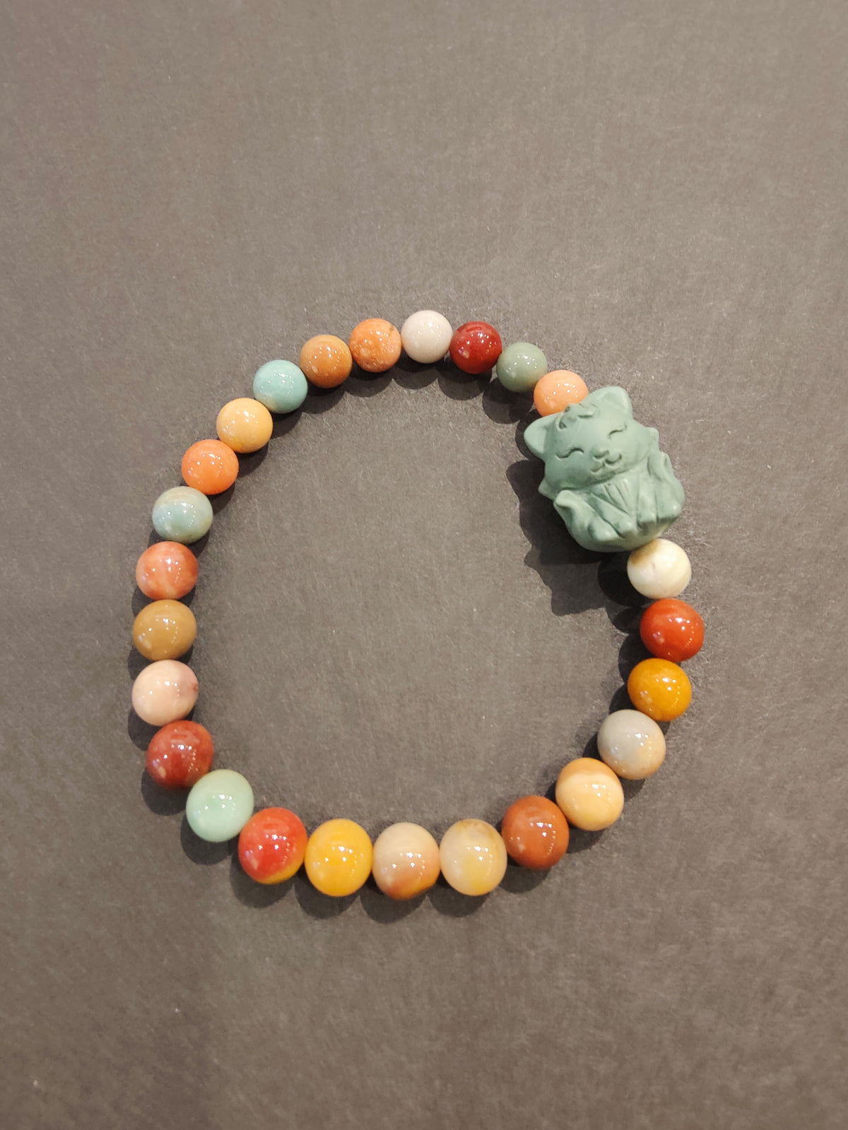 Beaded bracelet with round jasper stones in various earthy colors and a light blue-green cat-shaped animal charm, set against a gray background.