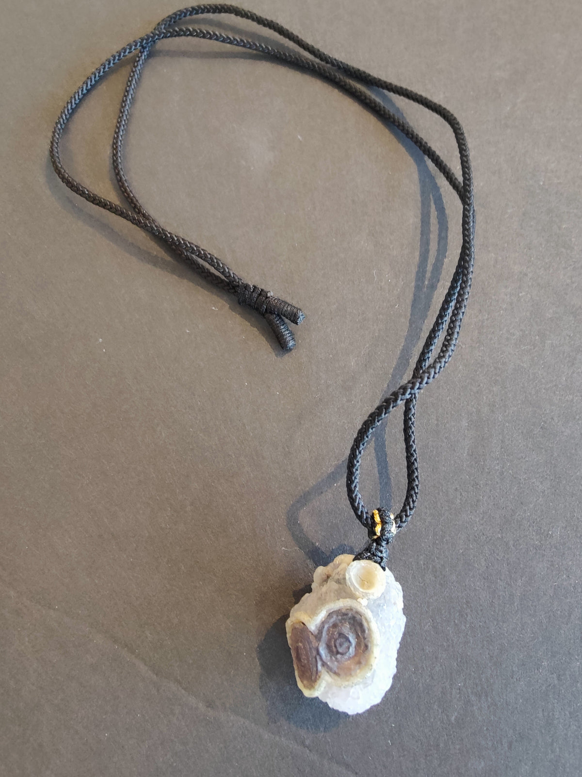 Earth-toned Gobi Desert eye agate pendant with circular patterns on a black braided cord necklace, displayed on a plain gray surface.