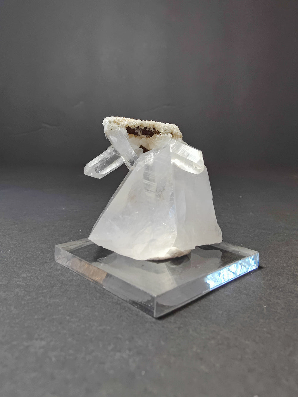 Cluster of clear quartz crystals anchored on a larger milky white quartz base, displayed upright on a transparent square stand against a dark gray background.