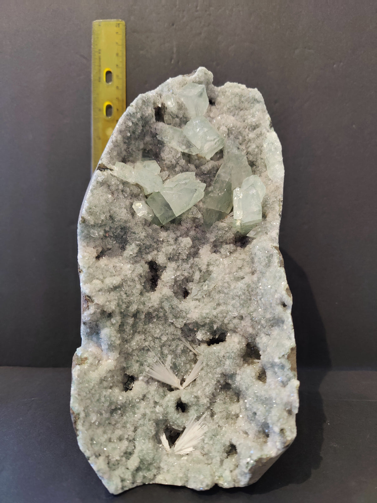 Gray and white crystalline apophyllite specimen with translucent, pale green rectangular crystals and feathery inclusions, displayed vertically next to a yellow measuring tool.