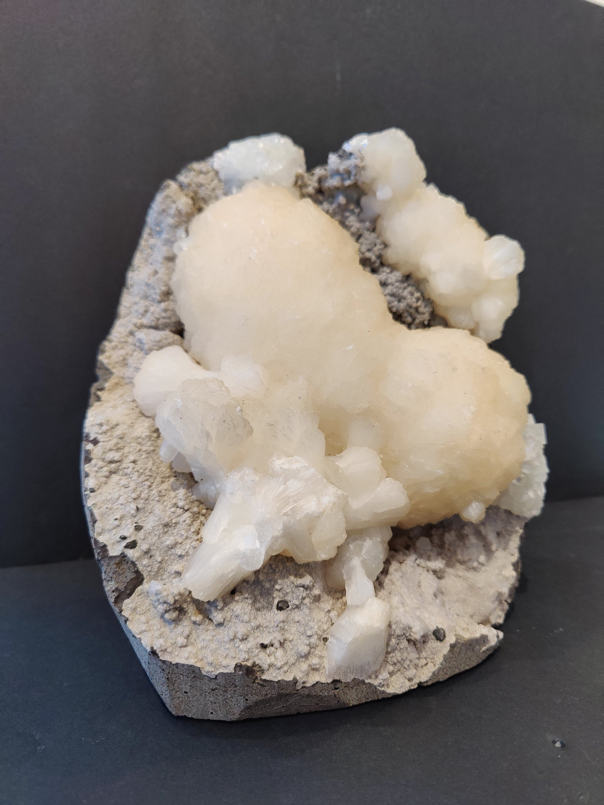 White botryoidal stilbite crystal formation resembling a heart atop a gray rocky matrix, accented by clear apophyllite crystals around the edges.
