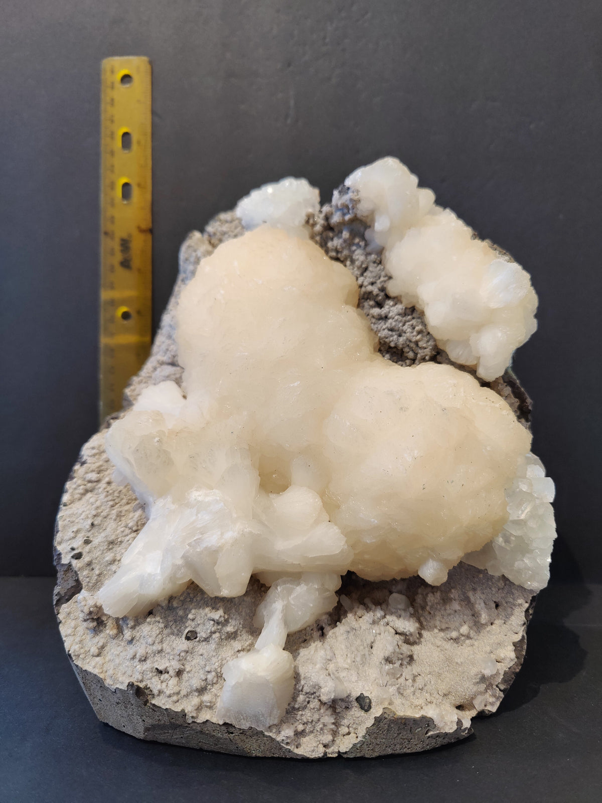 White Stilbite crystals clustered in a heart-like shape with clear Apophyllite crystals on a gray rough matrix, next to a yellow ruler for scale.