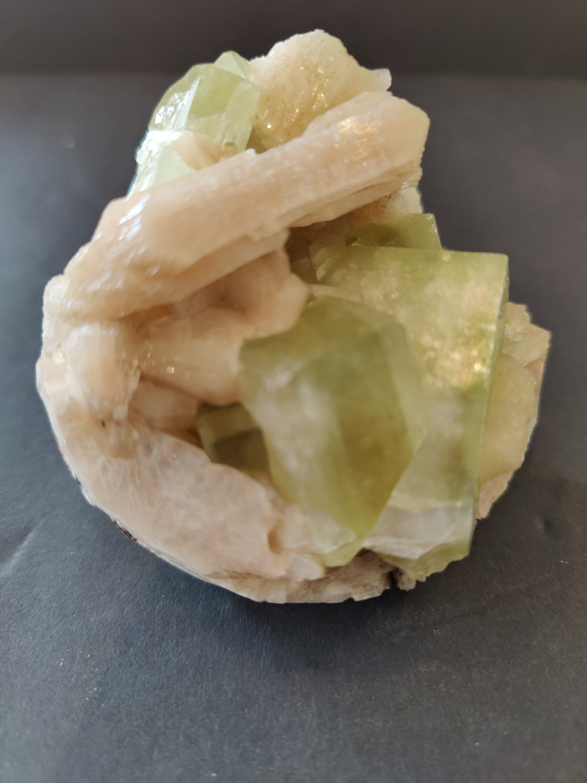 Cluster of pale peach stilbite crystals intergrown with translucent green apophyllite cubes on a dark gray surface, showing rough cubic and elongated shapes.
