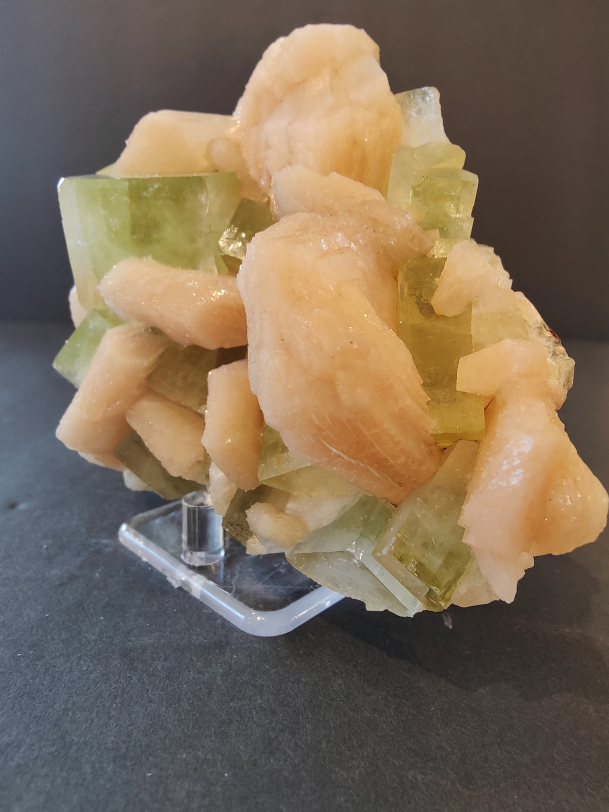 Cluster of translucent green apophyllite cubes intergrown with smooth, peach-colored stilbite formations, displayed on a clear stand against a dark background.
