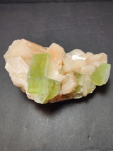 Cluster of transparent light green apophyllite cubes nestled among soft peach stilbite crystals on a dark surface, showing angular and layered formations.