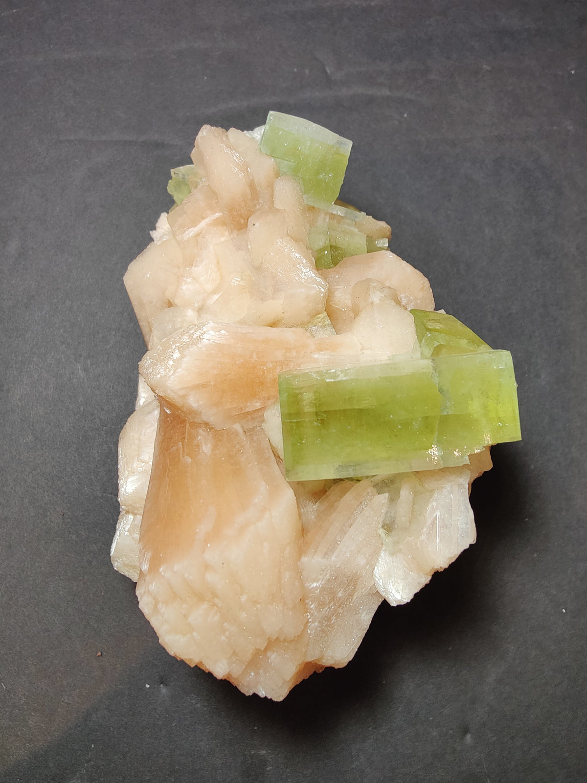 Cluster of bright green apophyllite cubic crystals resting on creamy peach stilbite formations against a dark surface background.