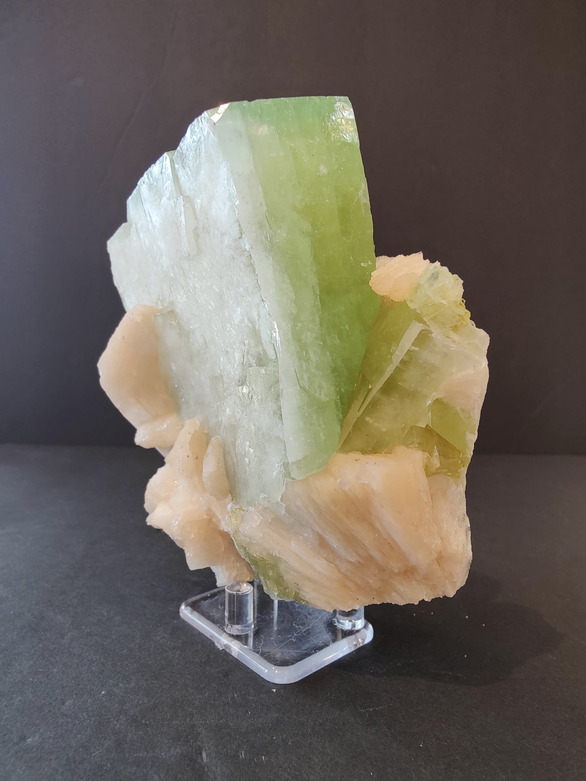 Light green translucent apophyllite crystal with peach-colored stilbite formations, standing on a clear acrylic display base against a dark background.
