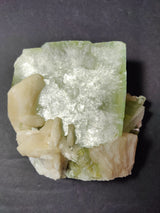 Square green Apophyllite crystal with translucent, reflective surface atop a cluster of peach-colored Stilbite crystals against a dark background.