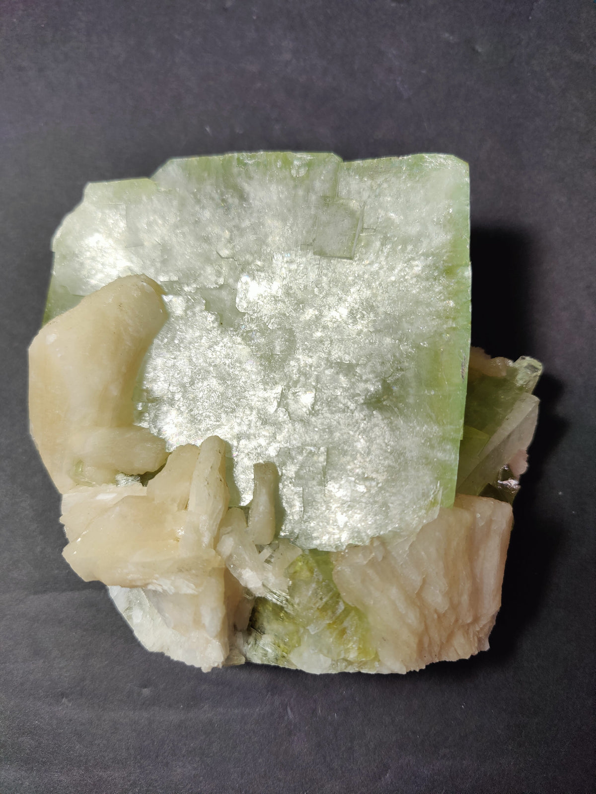 Square green Apophyllite crystal with translucent, reflective surface atop a cluster of peach-colored Stilbite crystals against a dark background.