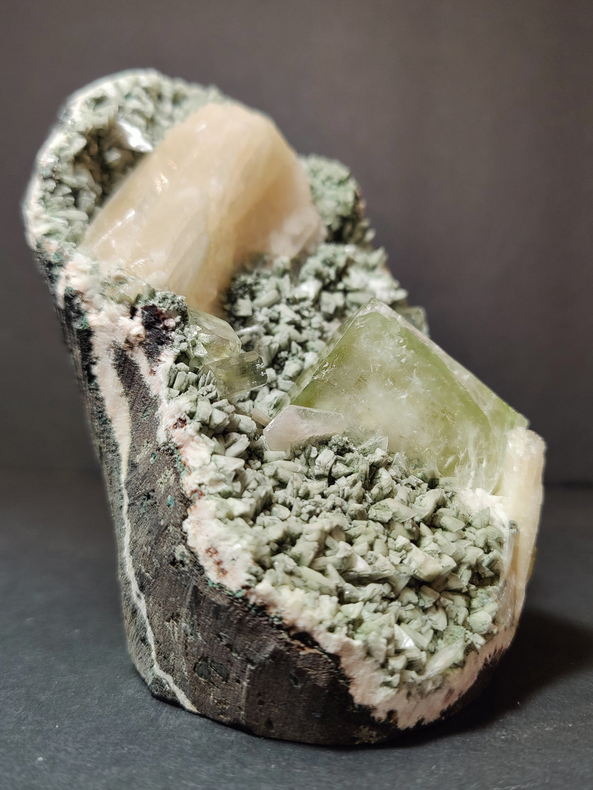 Cluster of pale green apophyllite cubes and peach stilbite crystals on a dark, rough heulandite base, viewed at a slight angle against a neutral background.