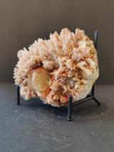 Cluster of pale pinkish-beige, needle-like stilbite crystals surrounding a rounded, cream-toned nodular formation, displayed on a black metal stand against a dark background.