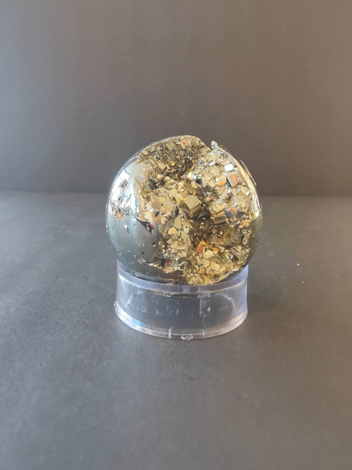 Polished pyrite sphere with metallic gold and silvery facets, displayed on a clear cylindrical stand against a neutral gray background.