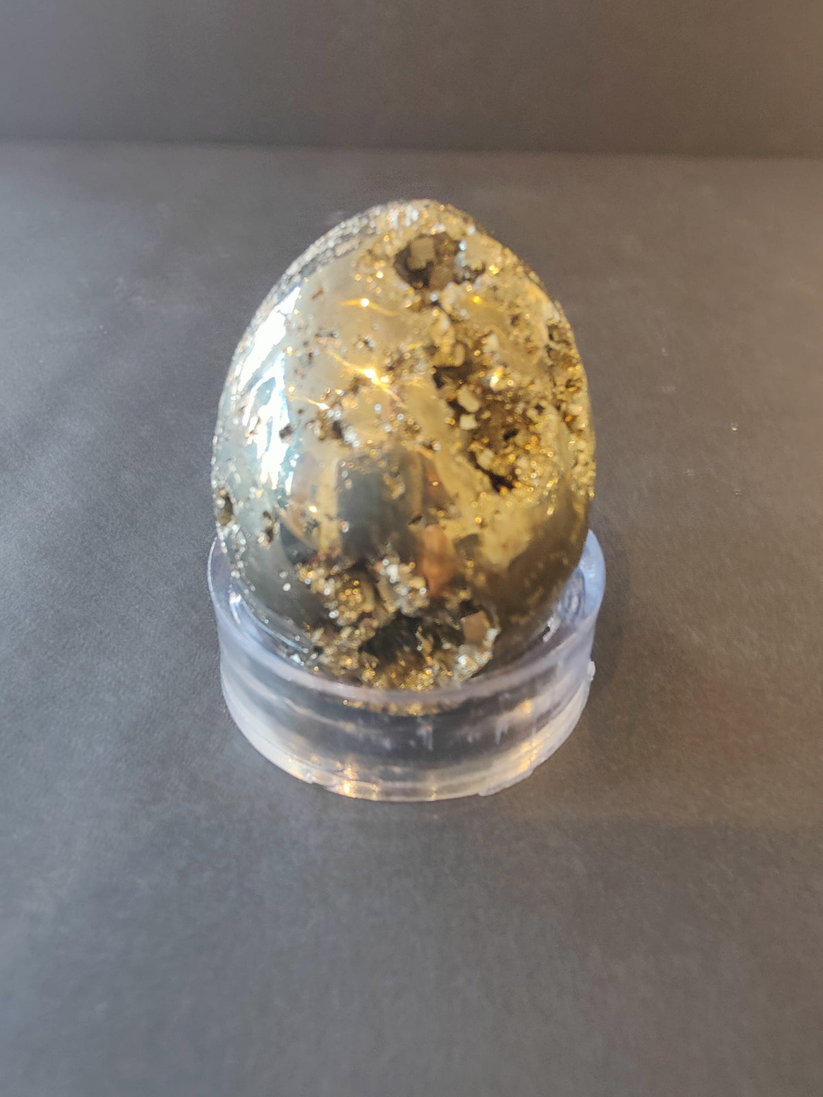 Polished egg-shaped pyrite stone with reflective gold and metallic patches, displayed on a clear circular stand against a muted background.