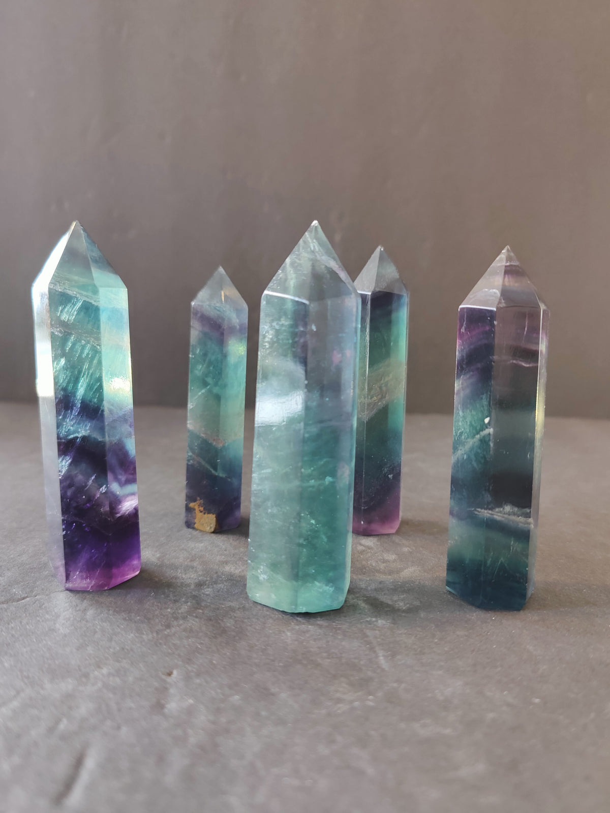 Five polished fluorite towers standing upright, showing bands of green, purple, and clear colors with pointed tops on a neutral surface and background.