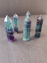 Five polished fluorite towers with pointed tops, displaying bands of green, purple, and blue hues, arranged standing on a neutral gray background.