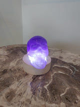Purple fluorite skull sculpture illuminated from beneath on a round wooden base, resting on a marbled stone surface against a plain background.