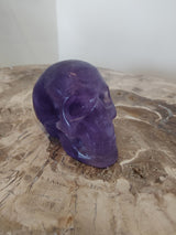 Smooth purple fluorite skull sculpture resting on a textured natural wood surface with visible grain patterns