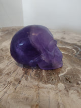 Translucent purple fluorite skull sculpture with smooth contours resting on a textured beige and brown stone surface, viewed from the side.