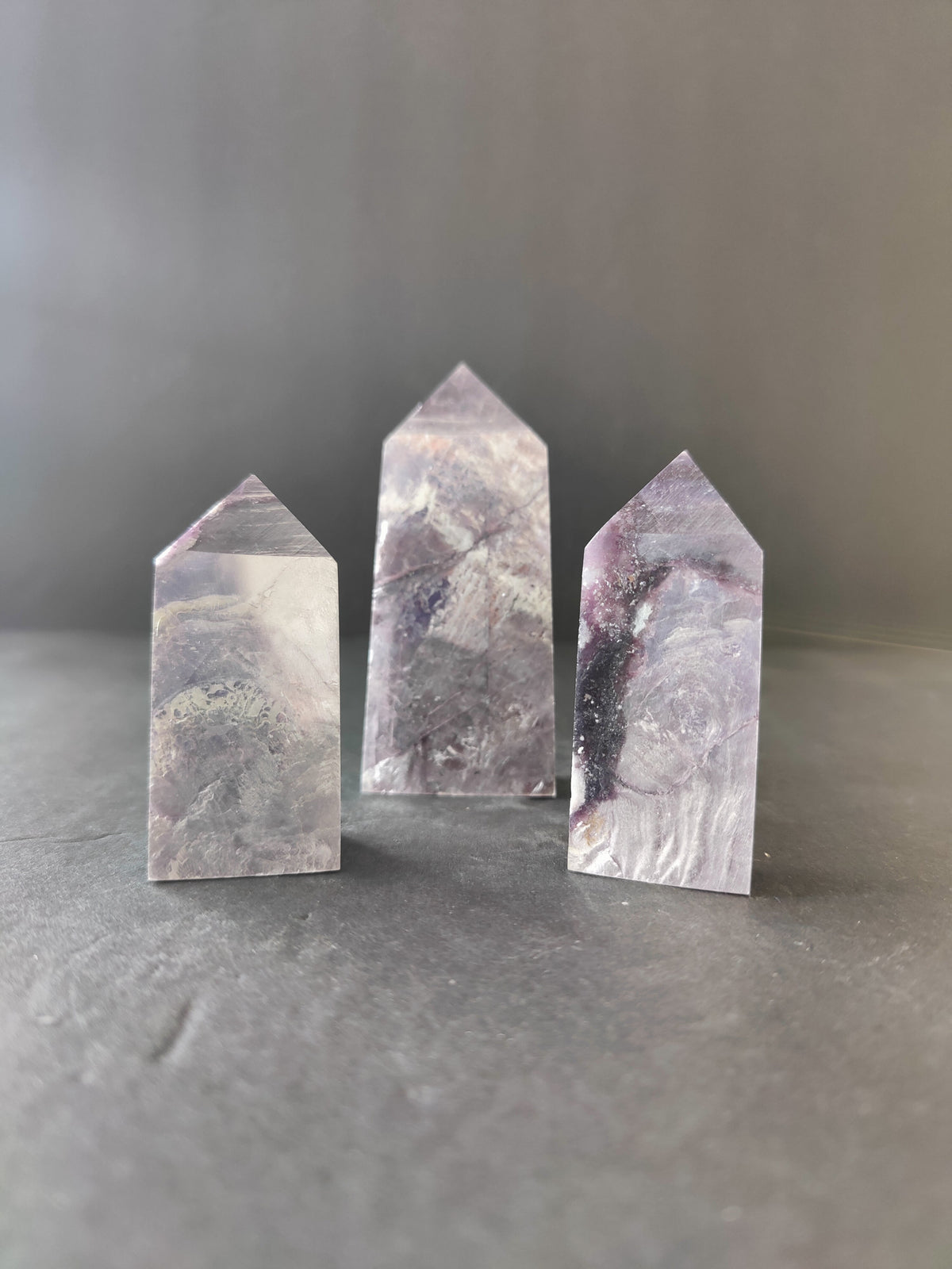 Three lepidolite tower sculptures with purple, lilac, and white marbled patterns, standing upright on a smooth gray surface with a soft gray background.