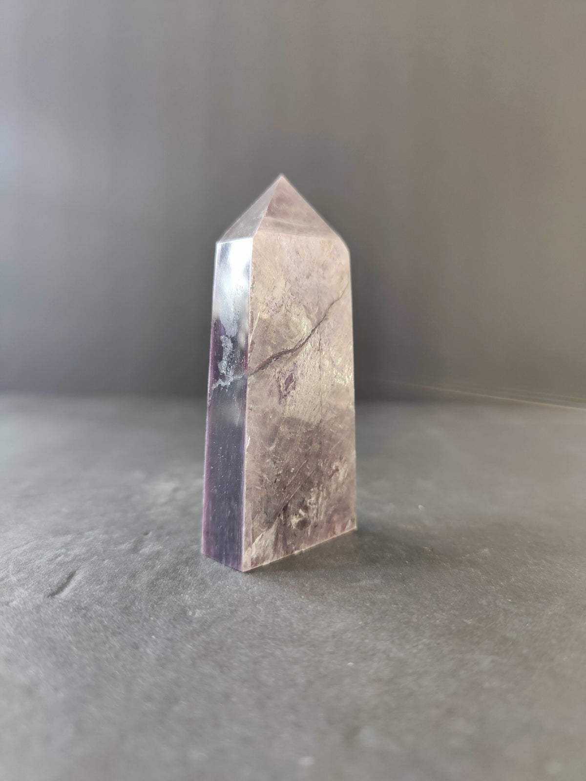 Translucent lepidolite tower sculpture with purple and beige hues, standing upright on a gray surface against a neutral gray background.