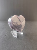 Polished heart-shaped lepidolite stone with lavender and white marbled patterns, displayed upright on a clear acrylic stand against a gray background.
