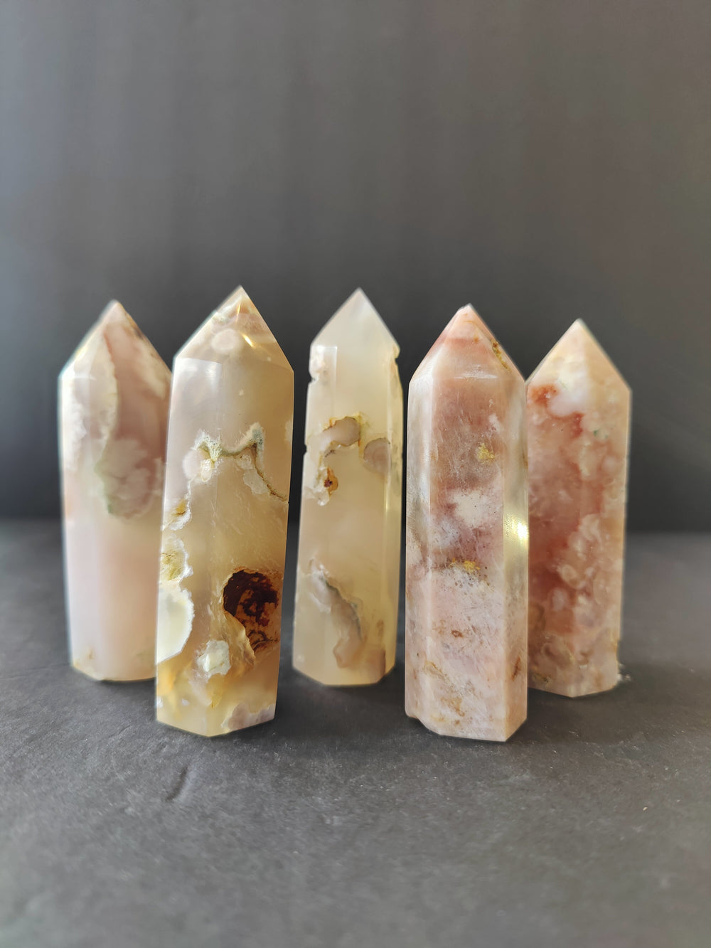 Cluster of five polished flower agate towers in soft pink, cream, and brown hues with natural patterns and pointed tops, arranged upright on a dark surface.