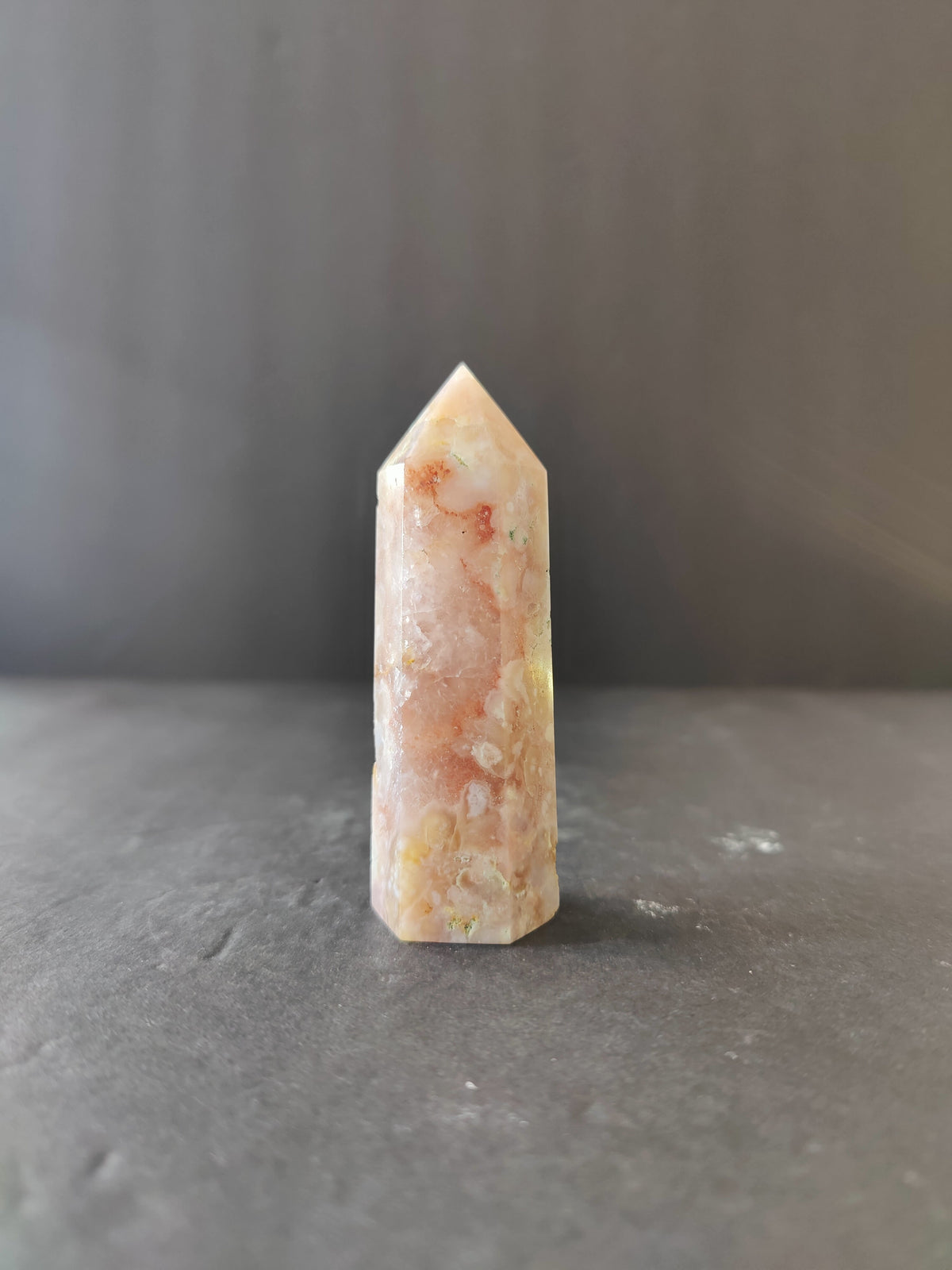 Tapered flower agate crystal tower with soft pink, white, and orange patterns, standing upright on a gray textured surface with a neutral backdrop.