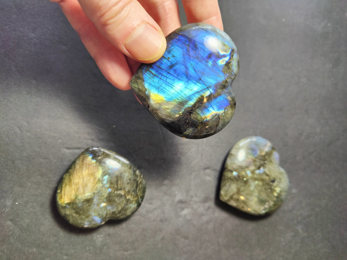Hand holding a blue-sheen labradorite heart-shaped polished stone above two similar heart stones with gold and gray iridescent flashes on a dark background.